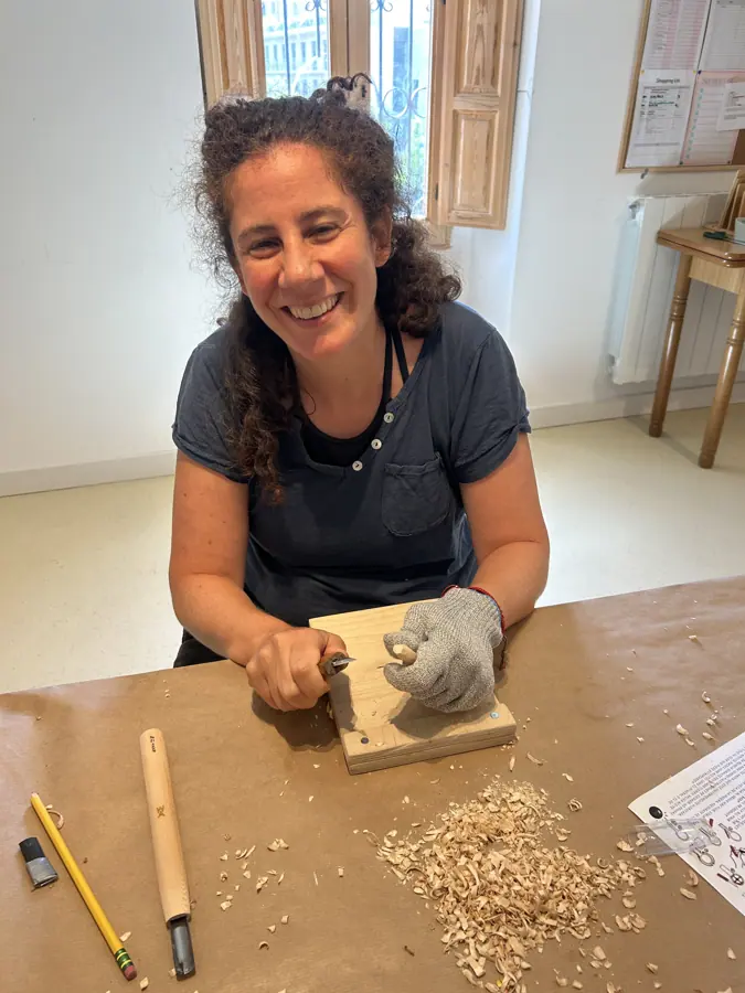 una participante sonriendo mientras talla una cuchara, con virutas sobre el banco